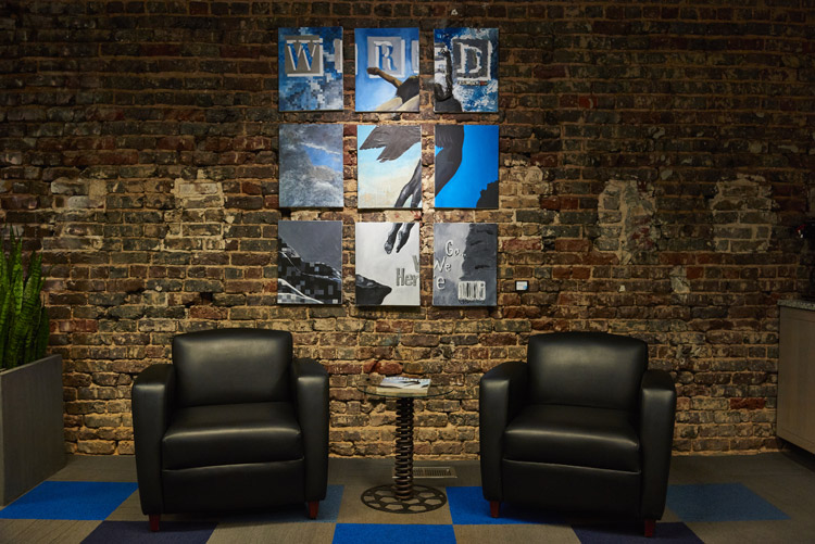 Photo of a section of Kinetic's conference room showcasing 2 chairs, a table made from train parts, and 9 paintings completing a recreation of the first issue of Wired magazine.