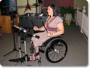 Photo of smiling woman in wheelchair playing Rockband with foosball table and tv behind her.